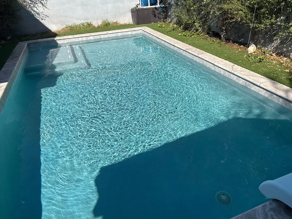Rectangular pool with sparkling turquoise water, submerged entry steps, light stone coping, and surrounding grass and vines, after pool service in Hawthorne, CA