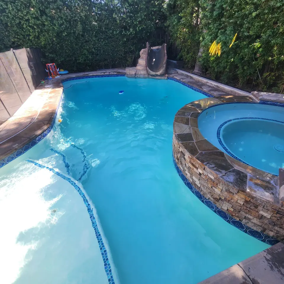 Clear blue pool with curved entry steps, a raised stone-clad spa, rock waterslide, and dense landscaping, after pool service in Hawthorne, CA