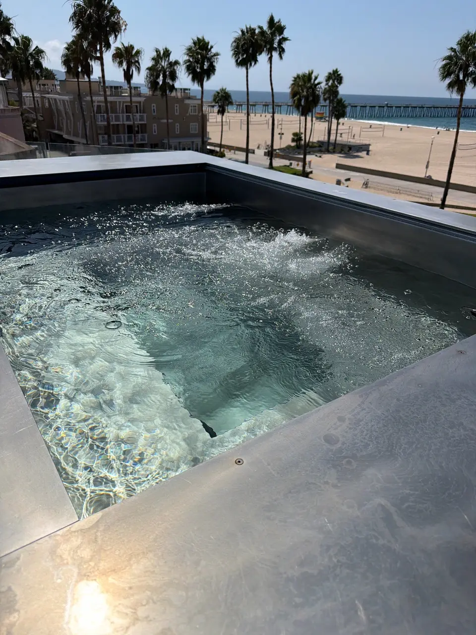 Clear water and bubbling jets fill a metal-surrounded spa overlooking a palm-lined beach and ocean, after pool service in Hermosa Beach, CA