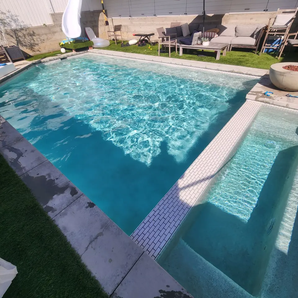 Sparkling turquoise water fills a rectangular pool and adjacent tiled spa, bordered by turf and patio seating, after pool service in Hermosa Beach, CA