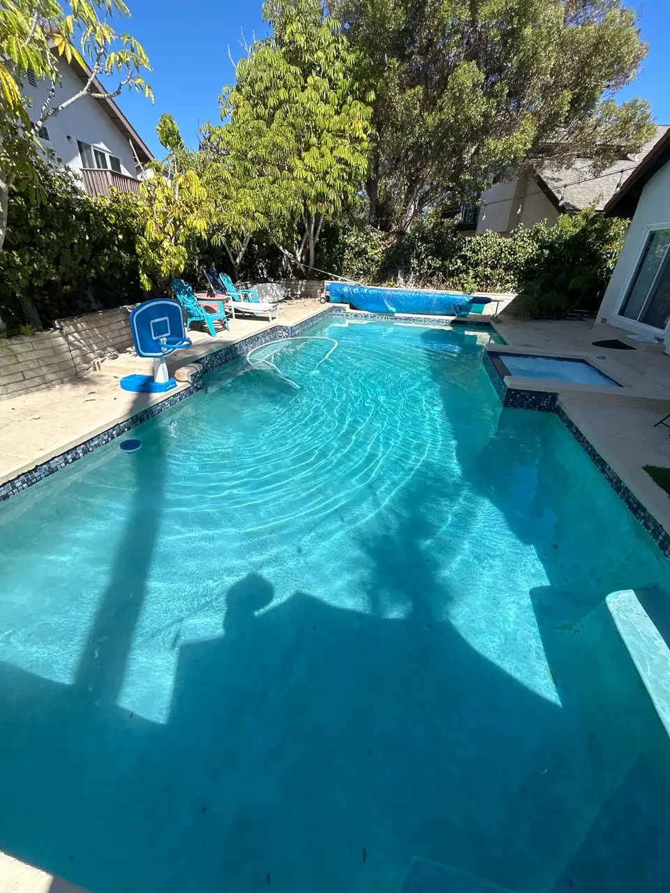 Sparkling turquoise pool with an attached spa, mosaic tile, basketball hoop, and surrounding trees, after pool service in La Jolla, CA