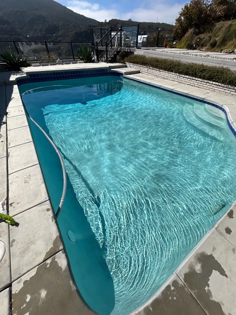 Sparkling turquoise pool water reveals curved steps beneath blue waterline tile, with glass fencing and hillside landscaping, after pool service in La Jolla, CA