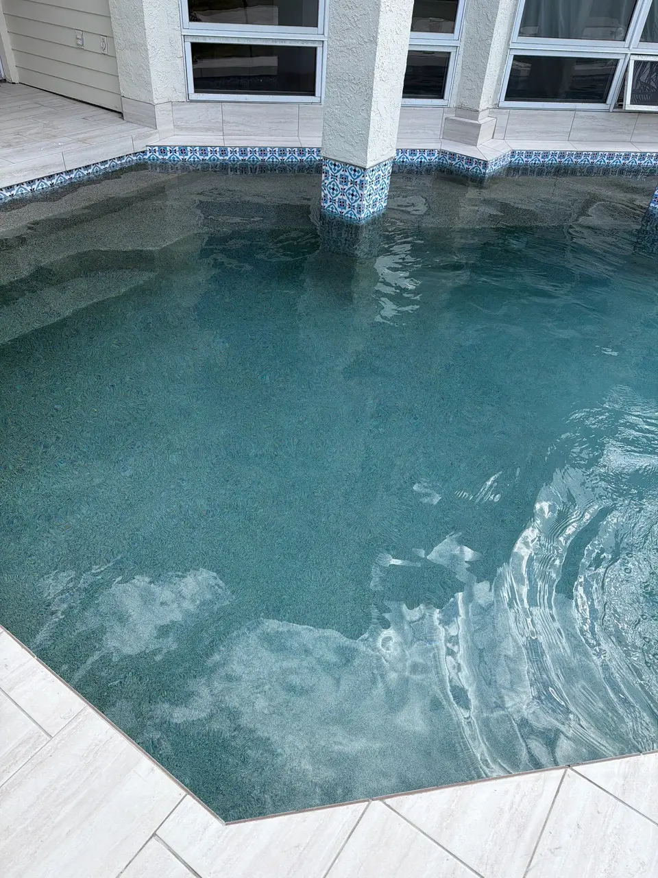 Clear blue pool water reveals a textured finish, submerged steps, and patterned tile around a support column, after pool service in La Jolla, CA