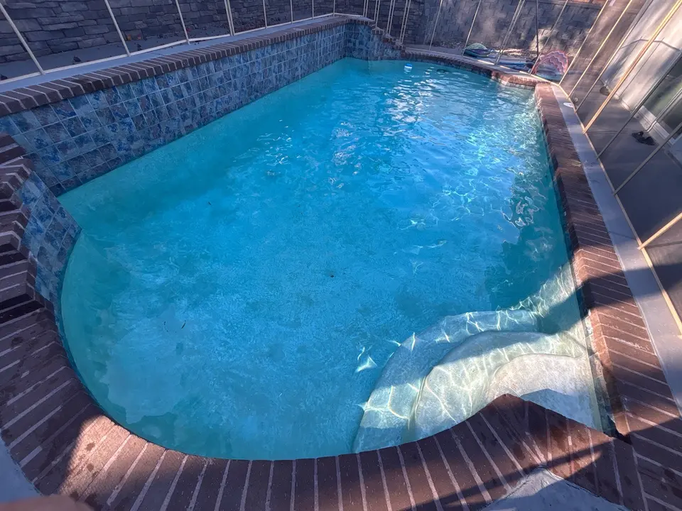 Clear blue pool water with curved entry steps, brick coping, and a raised blue-tiled wall inside a screen enclosure, after pool service in Lakeside, CA