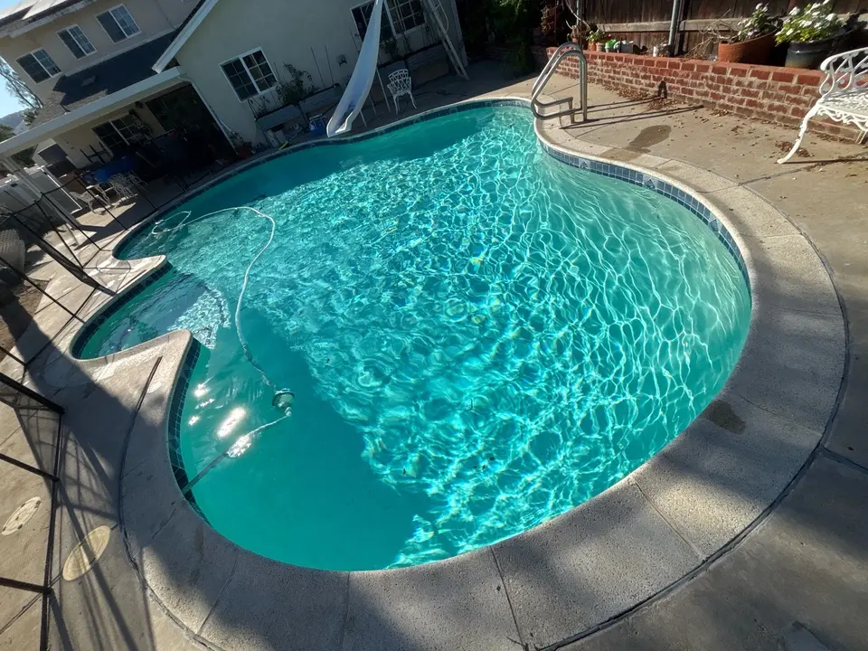 A curved pool has sparkling turquoise water, blue waterline tile, a slide, and a stainless steel handrail, after pool service in Lakeside, CA