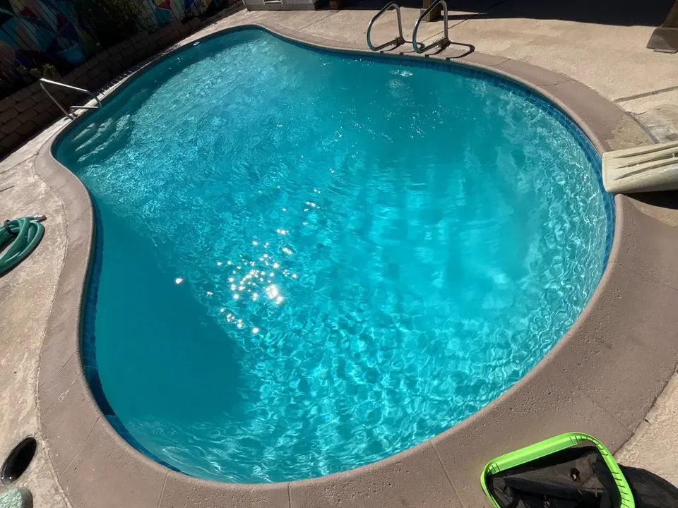 Freeform pool with sparkling turquoise water, blue waterline tile, entry steps, metal handrails, and a slide, after pool service in Lakeside, CA
