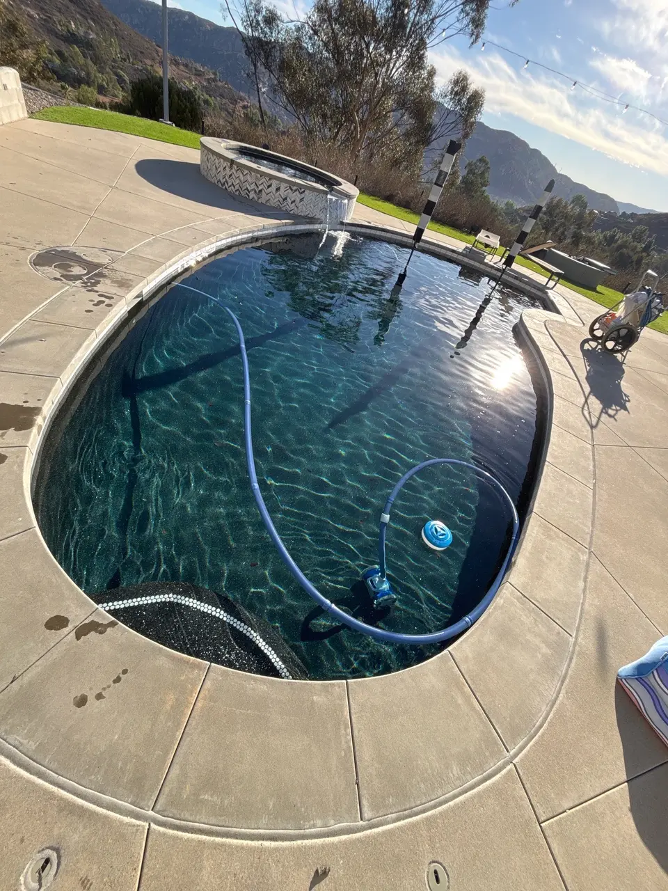 Clear dark teal water fills a curved pool with entry steps, a raised spillover spa, and mountain views, after pool service in Lakeside, CA