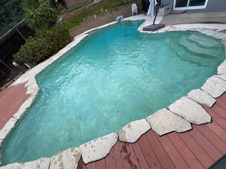 Clear turquoise water fills a freeform pool with submerged steps, stone coping, and a surrounding red deck, after pool service in Lakeside, CA