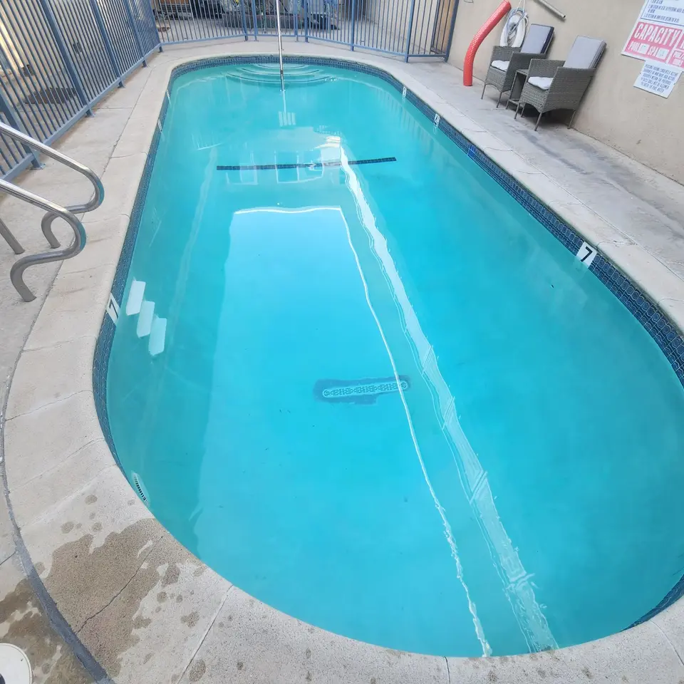 Clear turquoise pool with blue waterline tile, entry steps, stainless steel handrails, and surrounding safety fencing, after pool service in Lawndale, CA