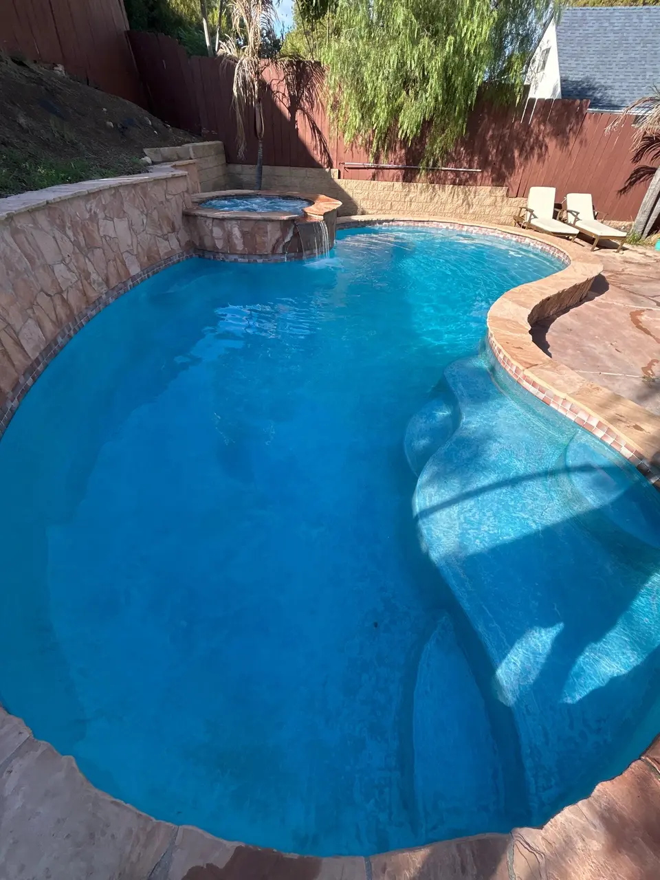Sparkling blue pool with curved entry steps, a raised spillover spa, stone walls, and surrounding landscaping, after pool service in Lemon Grove, CA