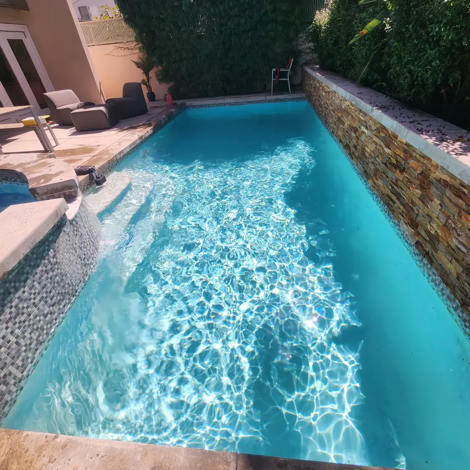 Sparkling blue pool water surrounds curved entry steps and a tiled spa beside a stacked-stone wall and greenery, after pool service in Manhattan Beach, CA