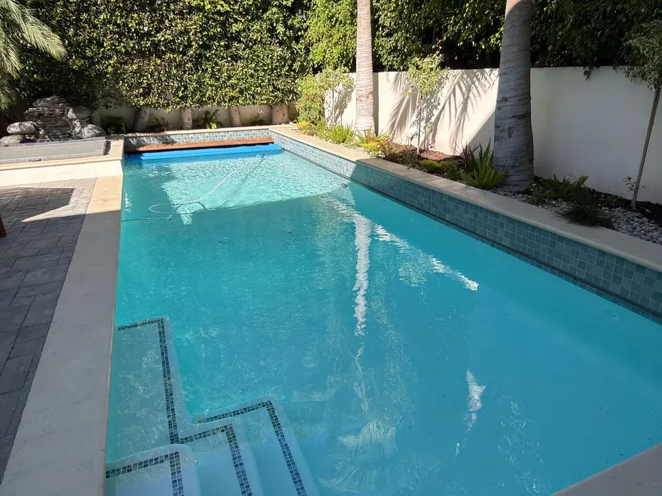 Clear turquoise water fills a rectangular pool with tiled steps, a stone waterfall, and surrounding palms and hedges, after pool service in Manhattan Beach, CA