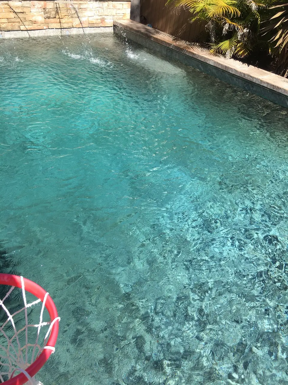 Clear turquoise pool water ripples beneath stone-wall water jets, with palms and a partially visible basketball hoop, after pool service in Manhattan Beach, CA