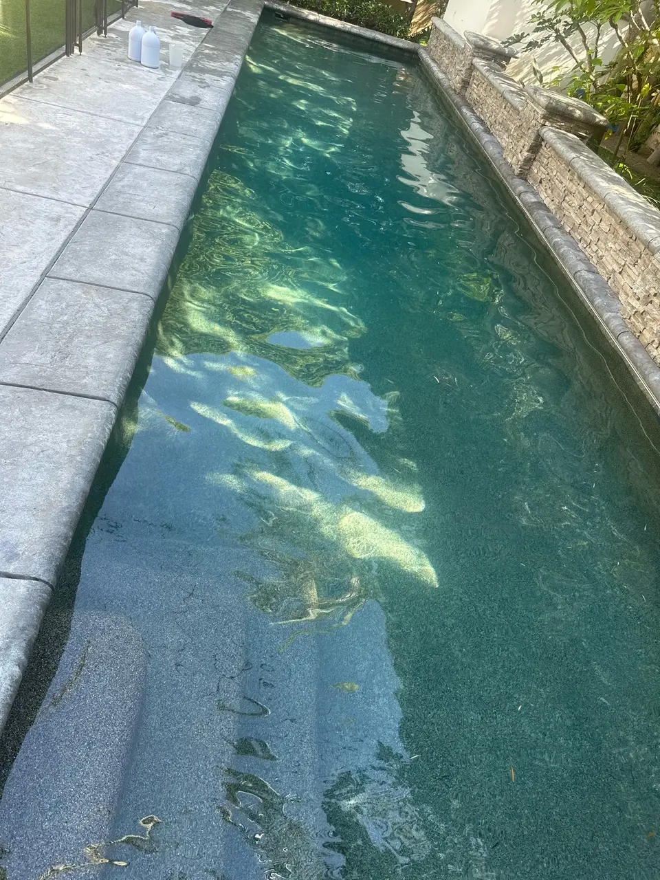 Clear teal water fills a narrow pool beside a stacked-stone wall with spillways and landscaping, after pool service in Manhattan Beach, CA