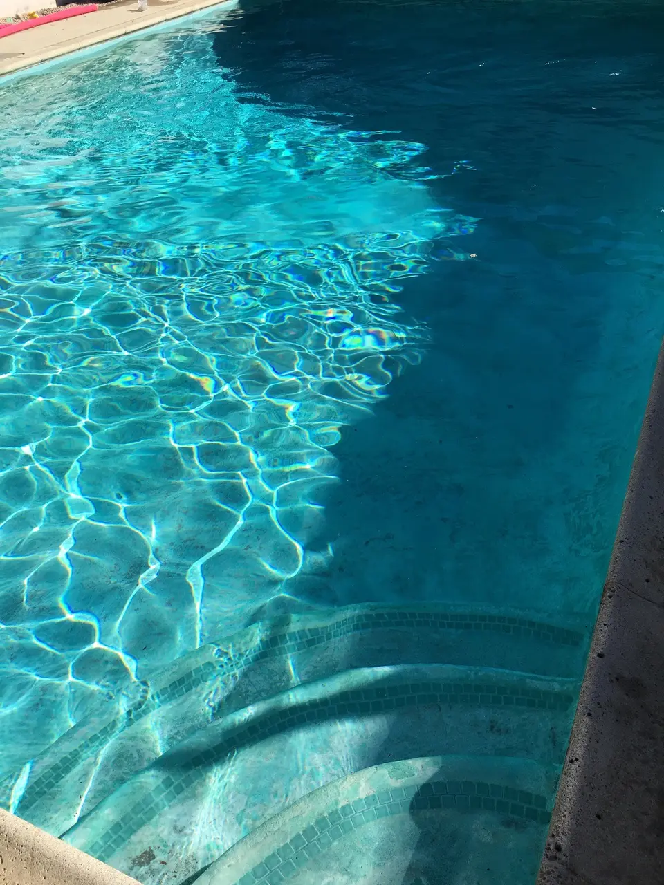 Clear blue water reveals curved, tile-bordered entry steps and a mottled pool finish beneath sunlight reflections, after pool service in Manhattan Beach, CA