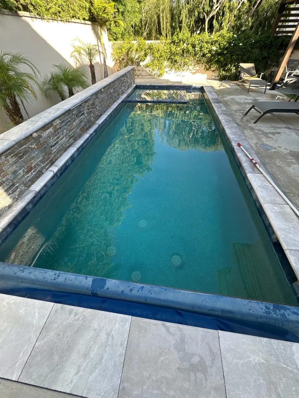 Clear turquoise water fills a rectangular pool with a spa, stone wall, submerged steps, and surrounding palms, after pool service in Manhattan Beach, CA