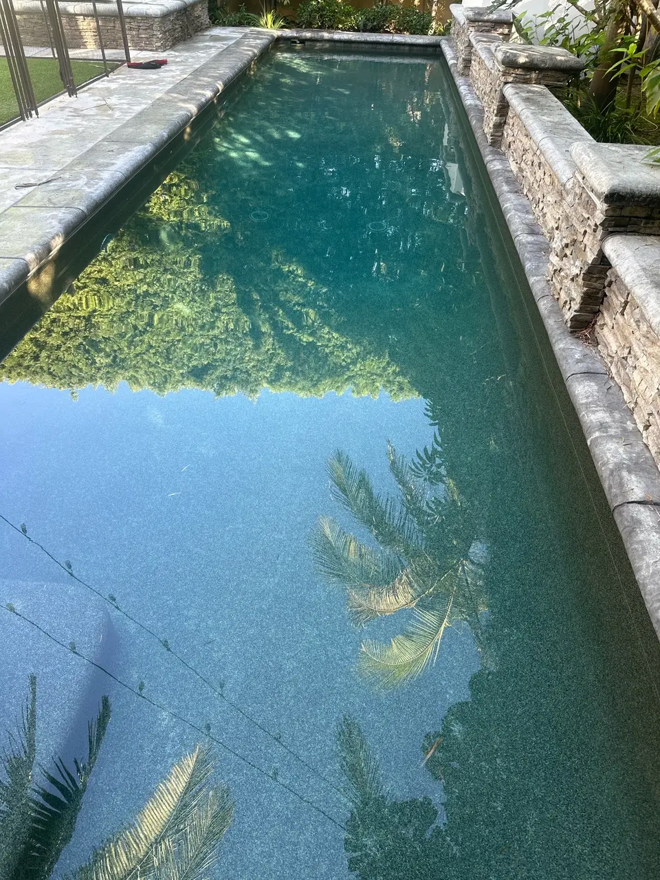 Clear deep teal water fills a narrow rectangular pool bordered by stone coping, stacked-stone walls, and tropical landscaping, after pool service in Manhattan Beach, CA