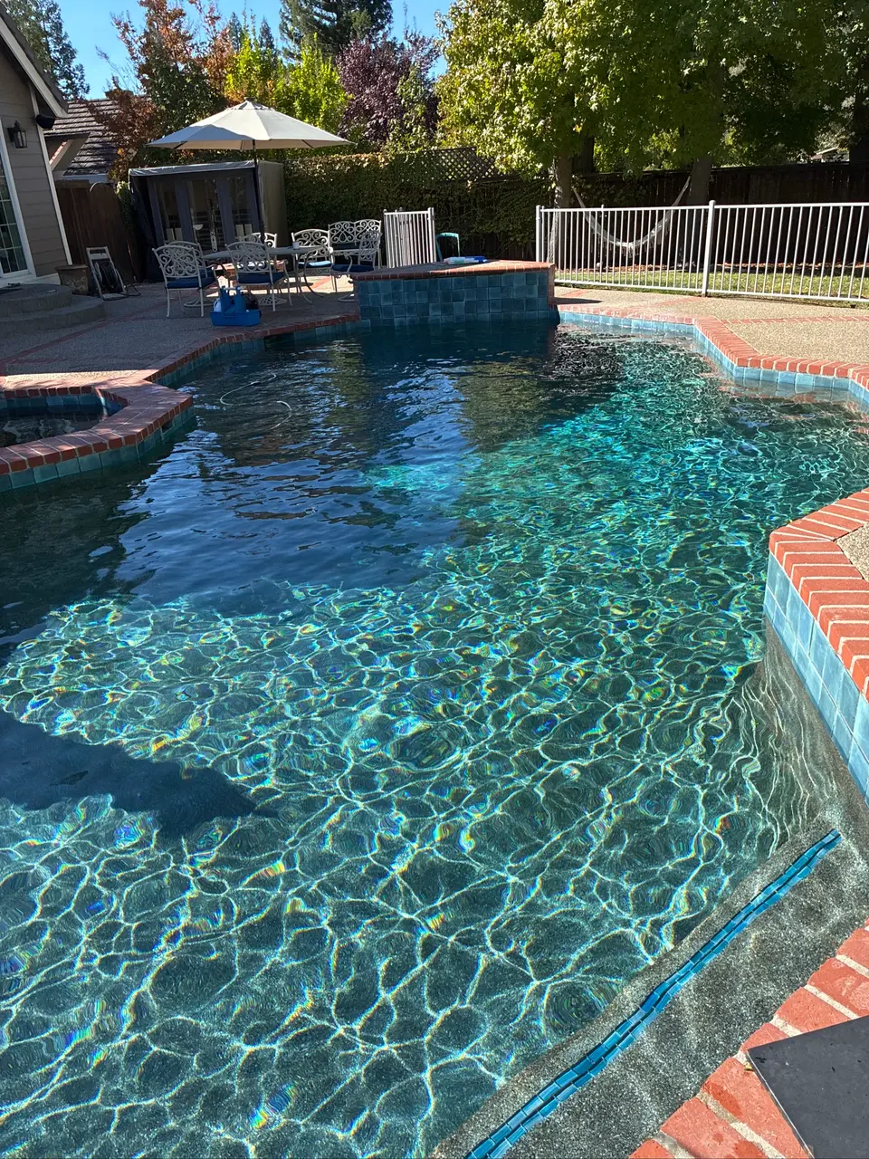 Sparkling blue pool water surrounds an attached spa, with brick coping, blue tile, and leafy landscaping, after pool service in Orangevale, CA