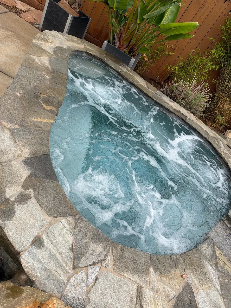 Clear blue spa water bubbles within natural stone coping, surrounded by tropical plants and a wooden fence, after pool service in Poway, CA