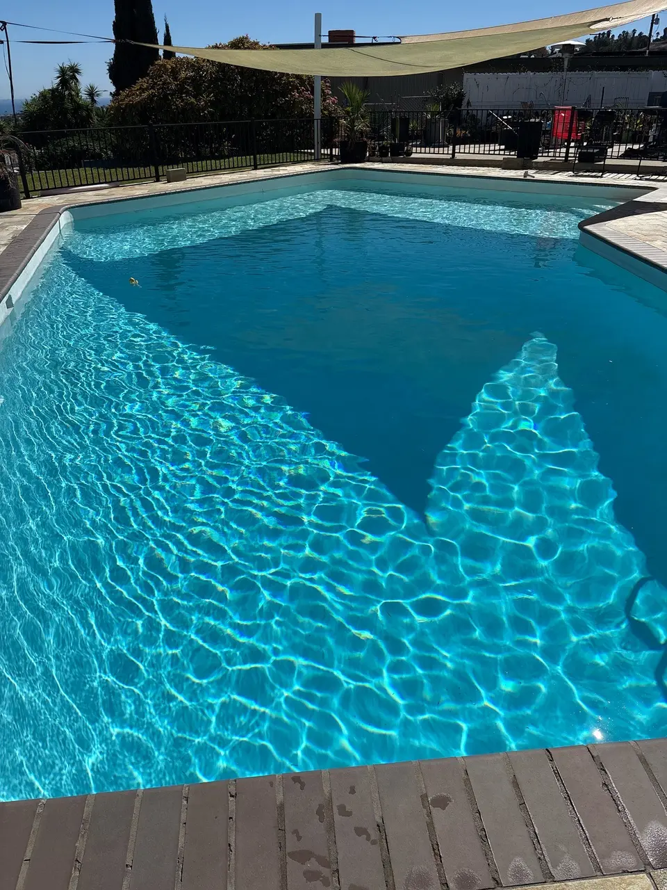Sparkling blue pool water reflects sunlight beneath a shade sail, surrounded by brick coping and a landscaped yard, after pool service in Rancho Palos Verdes, CA