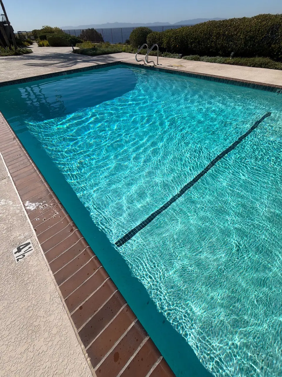 Sparkling turquoise pool water reveals a dark lane marking, bordered by brick coping, a ladder, and landscaped hedges, after pool service in Rancho Palos Verdes, CA