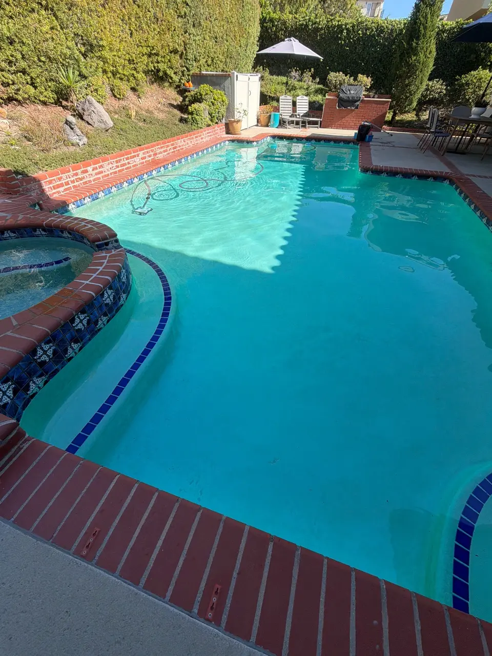 Clear turquoise pool water surrounds a raised tiled spa, with brick coping, curved steps, and tall hedges, after pool service in Rancho Palos Verdes, CA