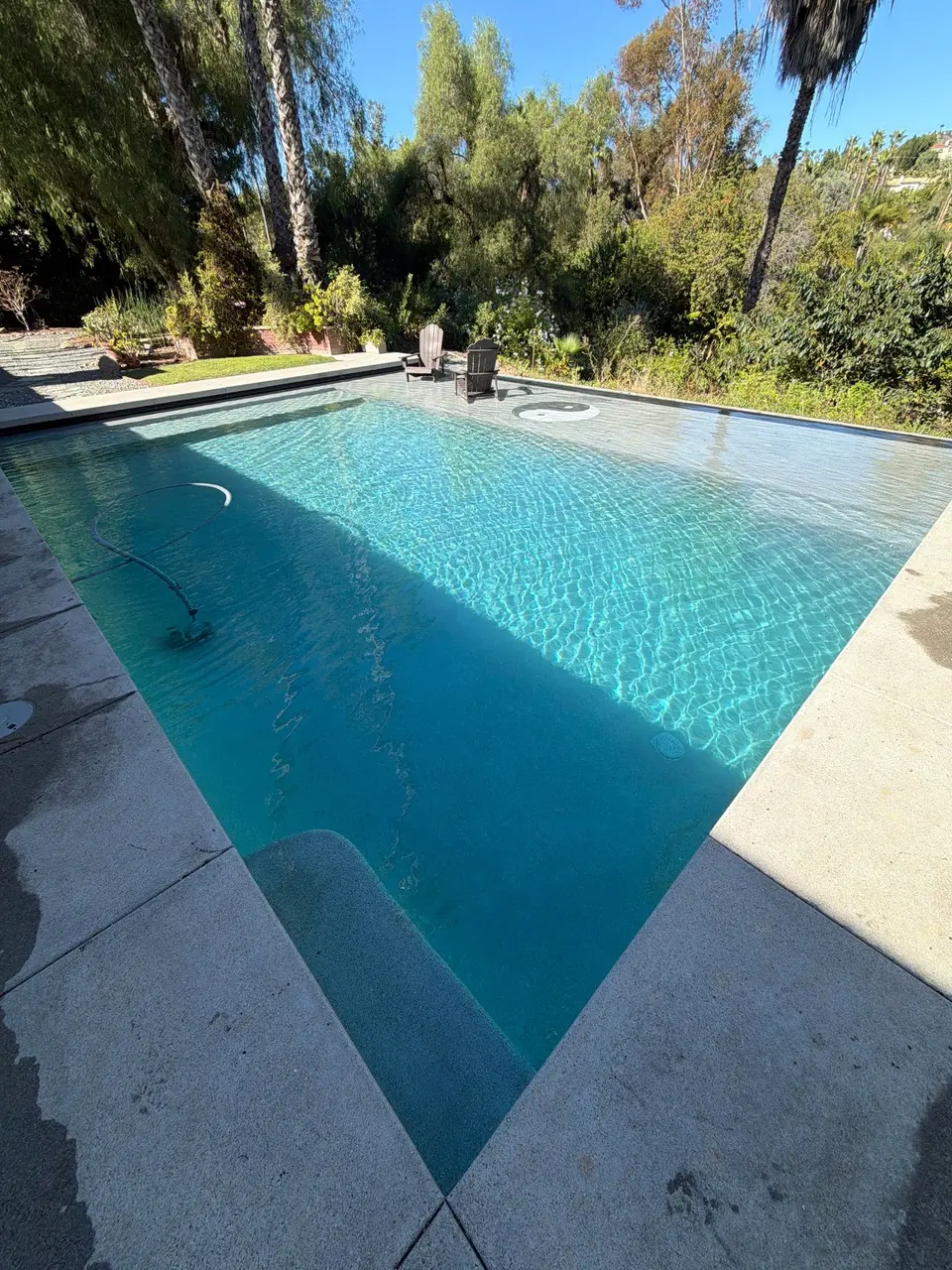 Sparkling turquoise pool with entry steps, a shallow sun shelf, in-water chairs, and lush surrounding trees, after pool service in Rancho Santa Fe, CA