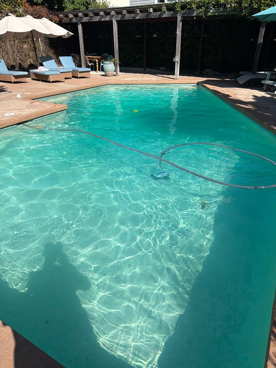 Clear turquoise pool water with submerged steps, a cleaner hose, and poolside loungers beneath umbrellas near a pergola, after pool service in Redondo Beach, CA