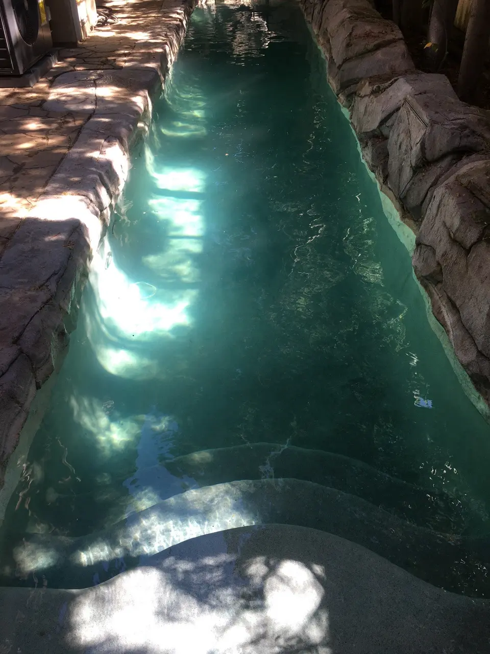 Clear teal water fills a narrow pool bordered by rock walls, stone decking, and curved entry steps, after pool service in Redondo Beach, CA
