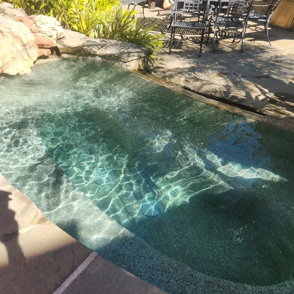 Clear water reveals pebble-finished spa seating beneath a rock waterfall, bordered by stone coping and landscaping, after pool service in Redondo Beach, CA