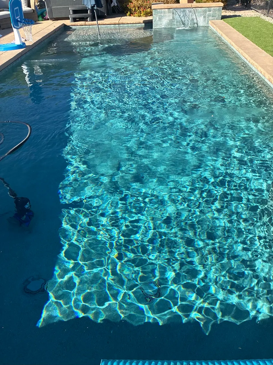 Sparkling blue pool water ripples beneath a tiled waterfall, with stone coping and a poolside basketball hoop, after pool service in Rocklin, CA