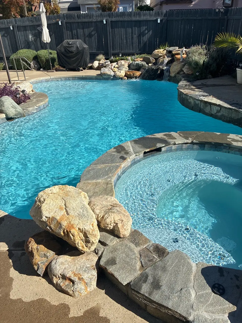 Sparkling turquoise pool and raised spa feature natural stone coping, a rock waterfall, and surrounding landscaping, after pool service in Rocklin, CA
