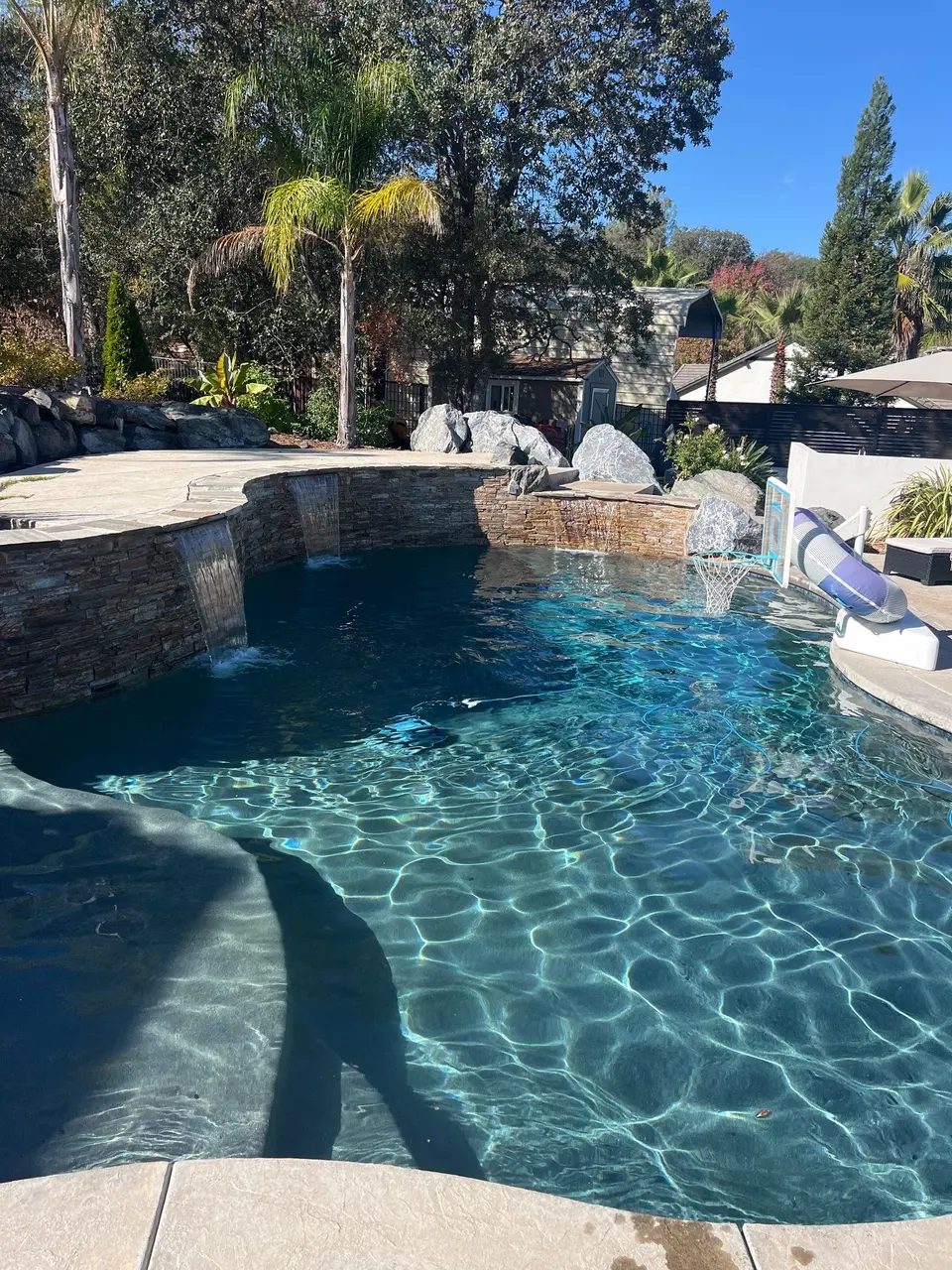 Sparkling blue pool with shallow steps, stone waterfalls, rock landscaping, a slide, and a basketball hoop, after pool service in Rocklin, CA