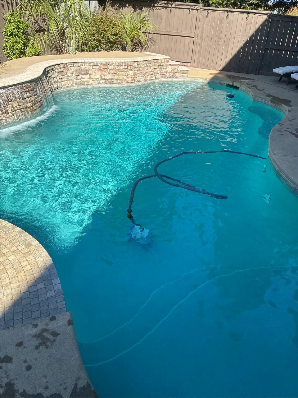 Clear turquoise pool water with submerged steps, a curved stone waterfall wall, and surrounding palms, after pool service in Rocklin, CA