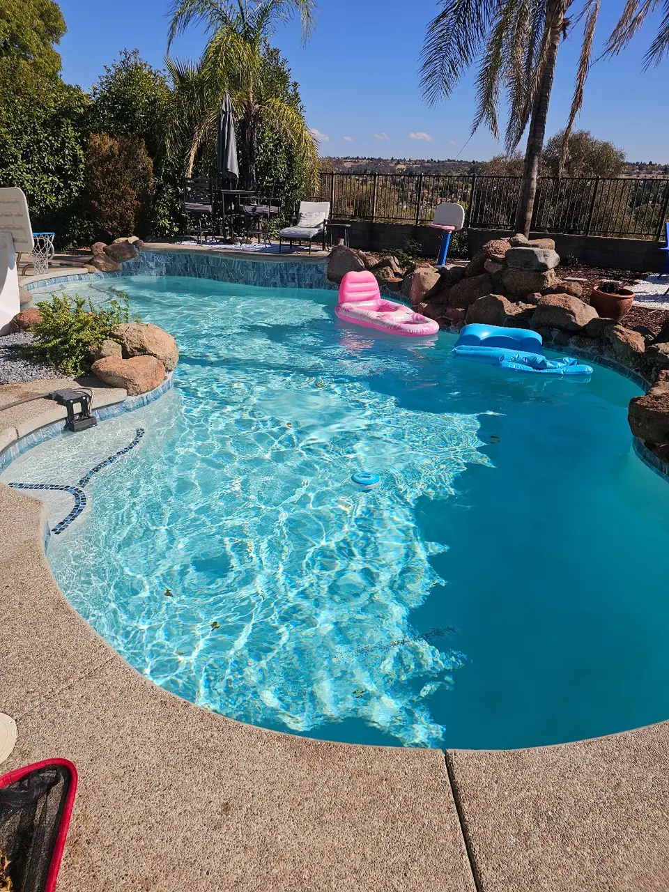 Clear turquoise pool water reveals shallow steps, surrounded by rock landscaping and palms, with colorful floats, after pool service in Rocklin, CA