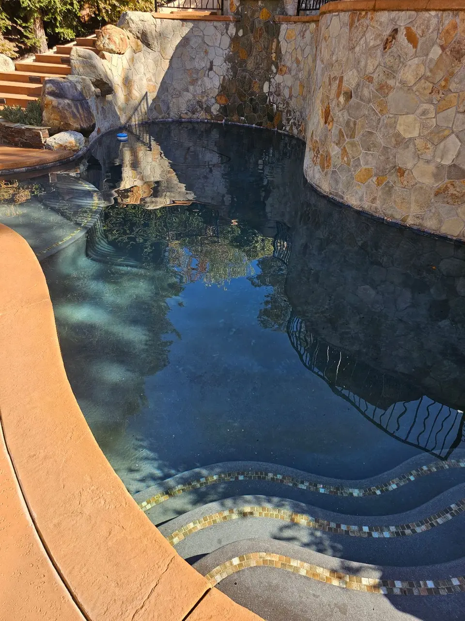 Clear dark-blue water fills a curved pool with tiled entry steps and tall stone walls, after pool service in Rocklin, CA