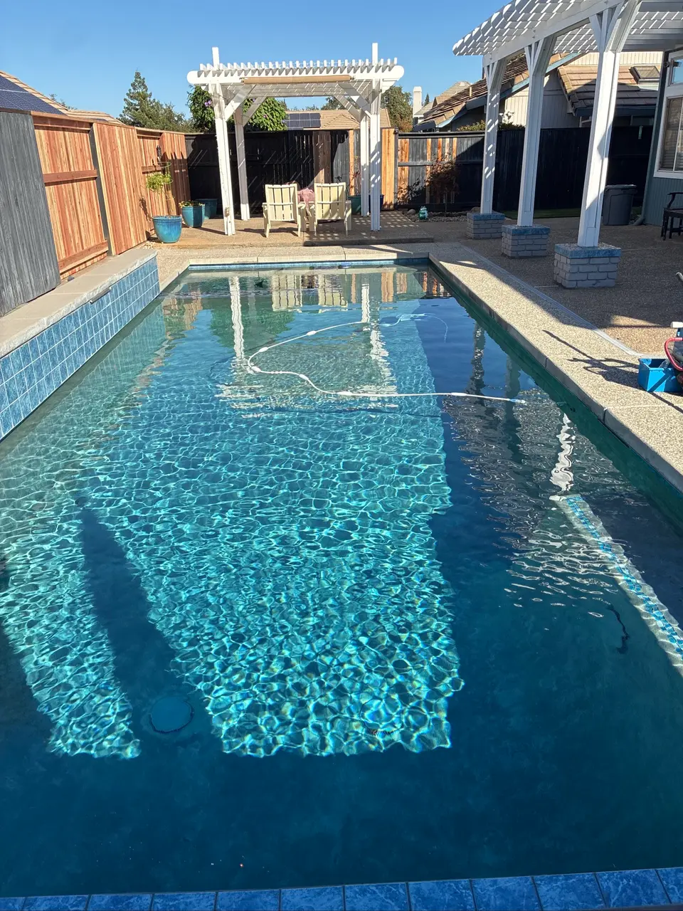 Rectangular pool with sparkling blue water, blue tile borders, a cleaner hose, and white pergolas beside poolside seating, after pool service in Rocklin, CA