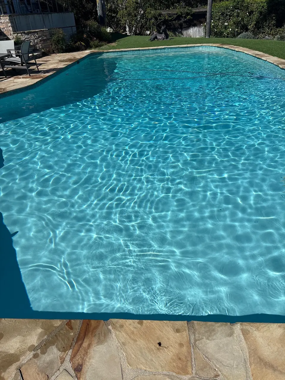 Sparkling turquoise pool water bordered by stone coping, waterline tile, lawn, and mature landscaping, after pool service in Rolling Hills, CA