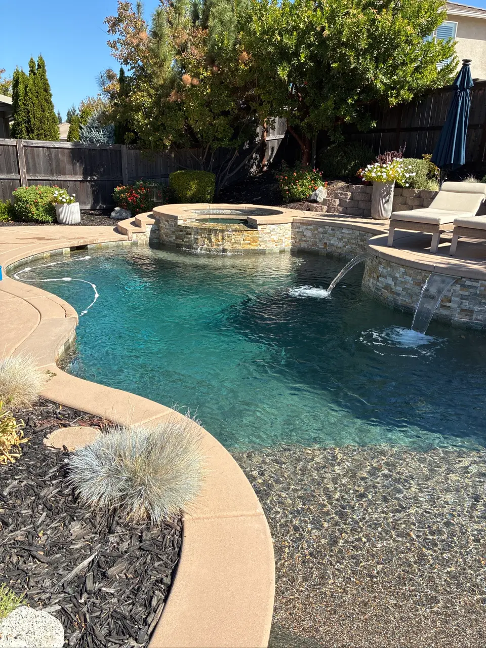 Sparkling turquoise pool with a raised spa, stone-faced waterfalls, pebble finish, and landscaped surroundings, after pool service in Roseville, CA
