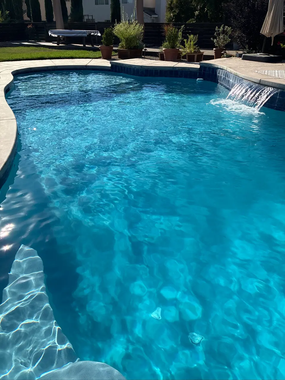 Sparkling blue pool water reveals curved entry steps beneath a tiled waterfall, with potted plants bordering the deck, after pool service in Roseville, CA