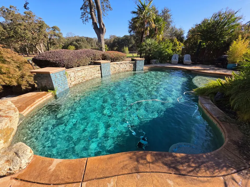 Sparkling turquoise water fills a freeform pool with blue tile, stacked-stone walls, rock edging, and lush landscaping, after pool service in Roseville, CA