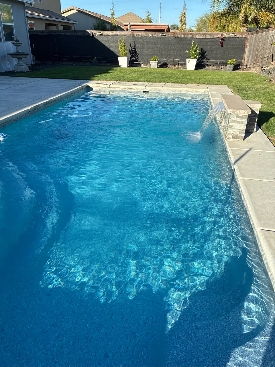 Sparkling blue pool water surrounds submerged steps beneath a raised stone waterfall, bordered by lawn and potted plants, after pool service in Roseville, CA