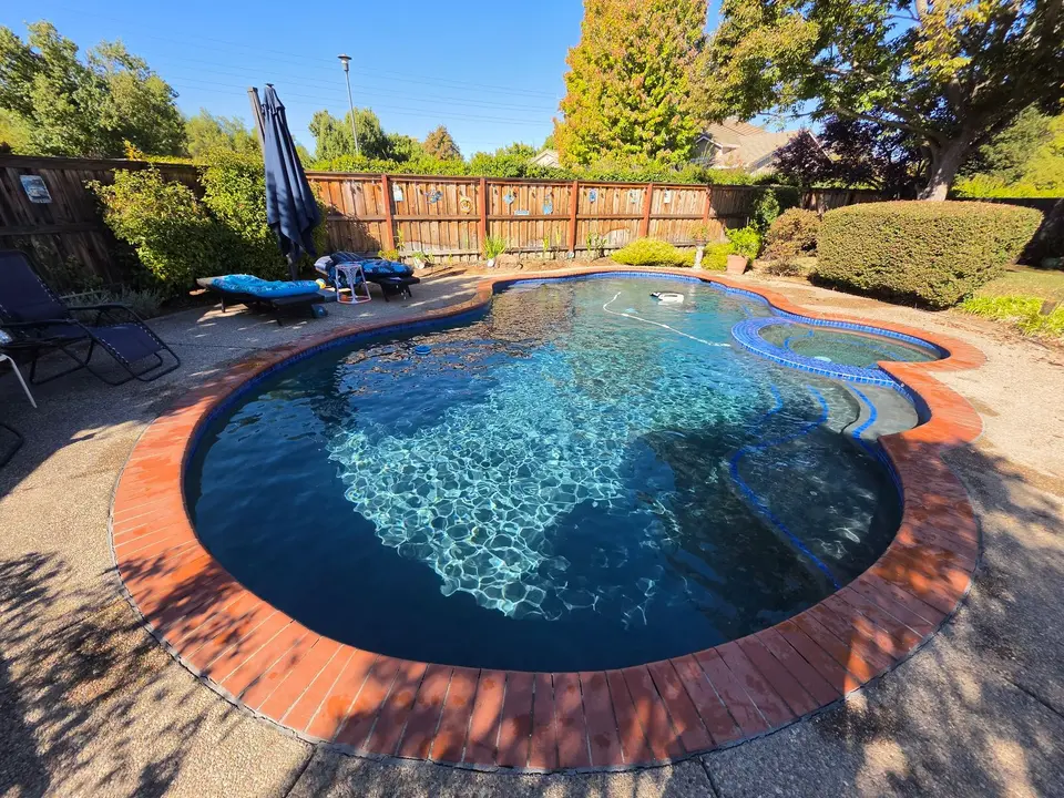 Sparkling blue water fills a freeform pool with an attached spa, curved steps, brick coping, and surrounding landscaping, after pool service in Roseville, CA