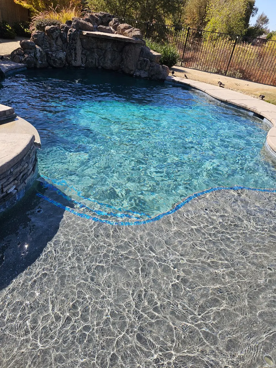 Sparkling blue pool water surrounds a shallow sun shelf with blue tile accents beneath a rock waterfall, after pool service in Roseville, CA