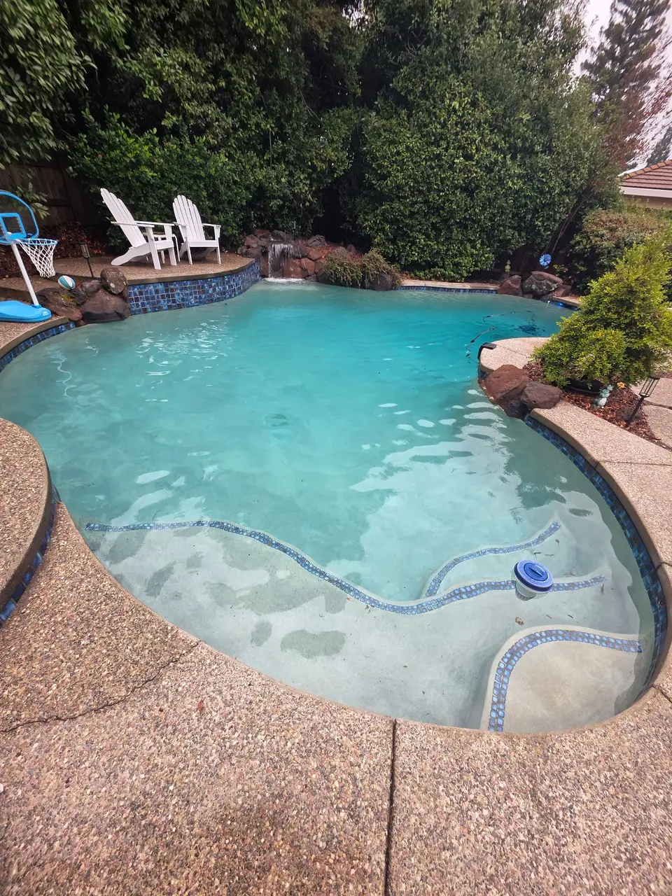 Clear turquoise water fills a freeform pool with tiled entry steps, a rock waterfall, and dense landscaping, after pool service in Roseville, CA