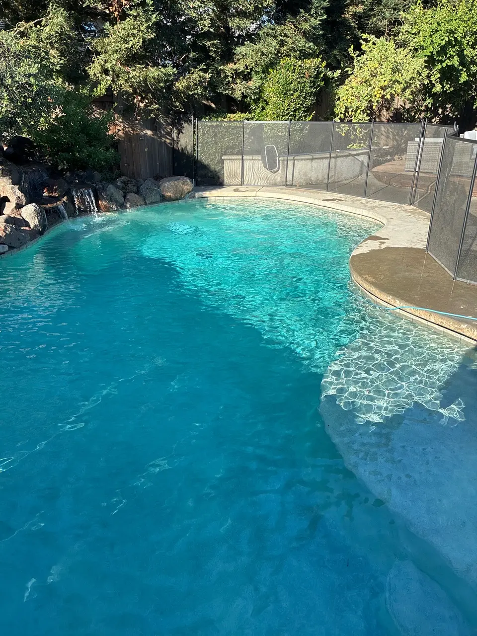 Sparkling turquoise water fills a curved pool with shallow steps, a rock waterfall, and surrounding greenery, after pool service in Roseville, CA