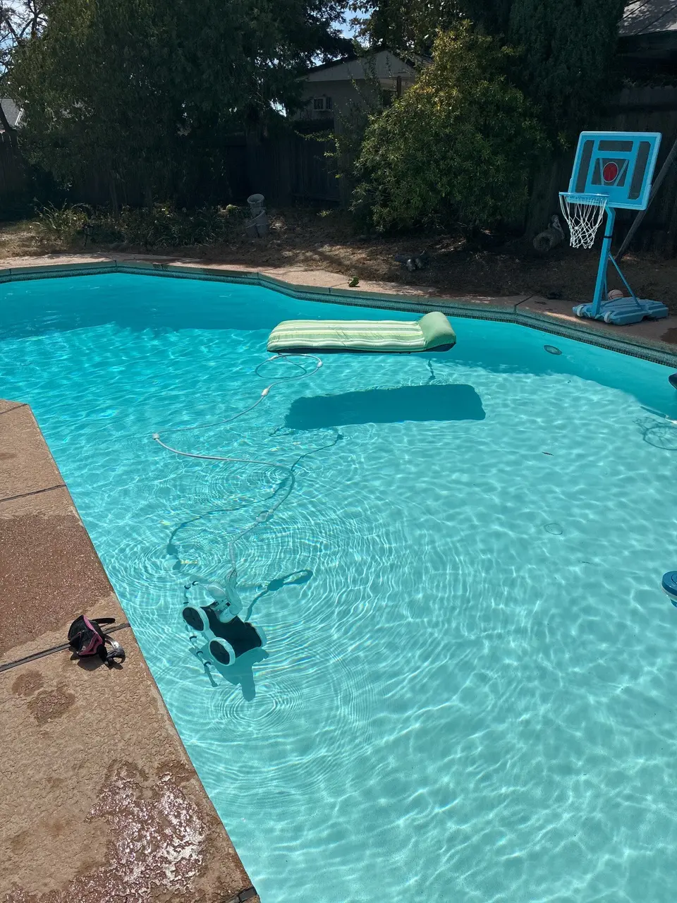 Clear turquoise pool water reveals a light-colored finish, robotic cleaner, floating lounge, and poolside basketball hoop, after pool service in Sacramento,, CA
