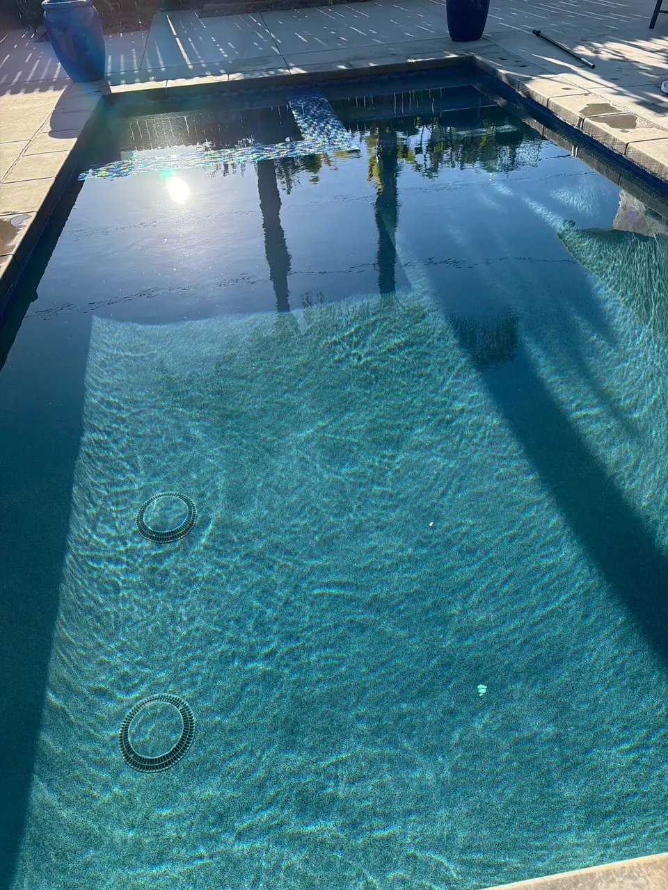 Clear blue water reveals a textured pool finish, mosaic tile accents, and submerged steps beneath tree reflections, after pool service in San Diego, CA