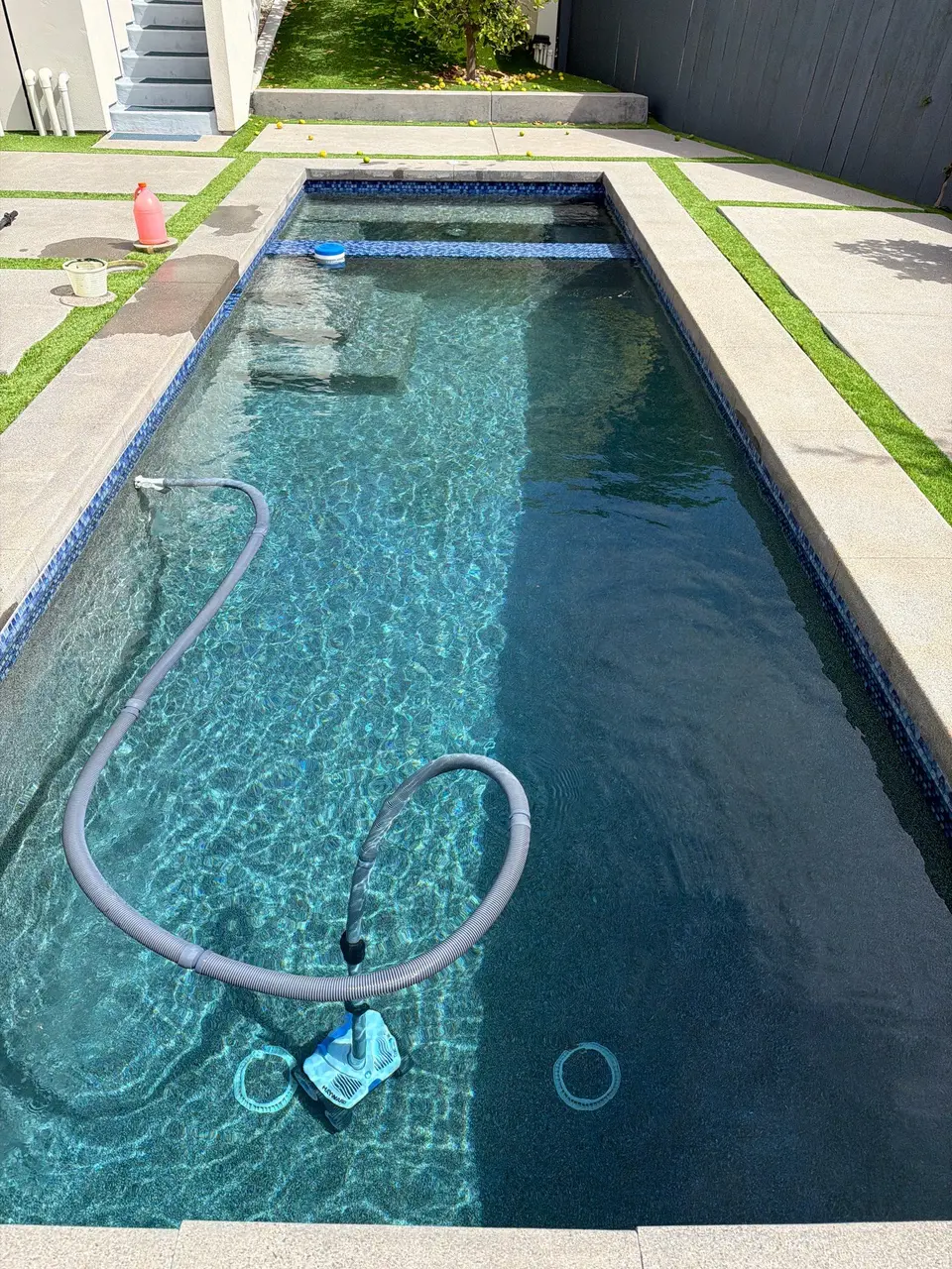 Clear turquoise water fills a narrow rectangular pool with blue mosaic tile, entry steps, and a submerged cleaner, after pool service in San Diego, CA