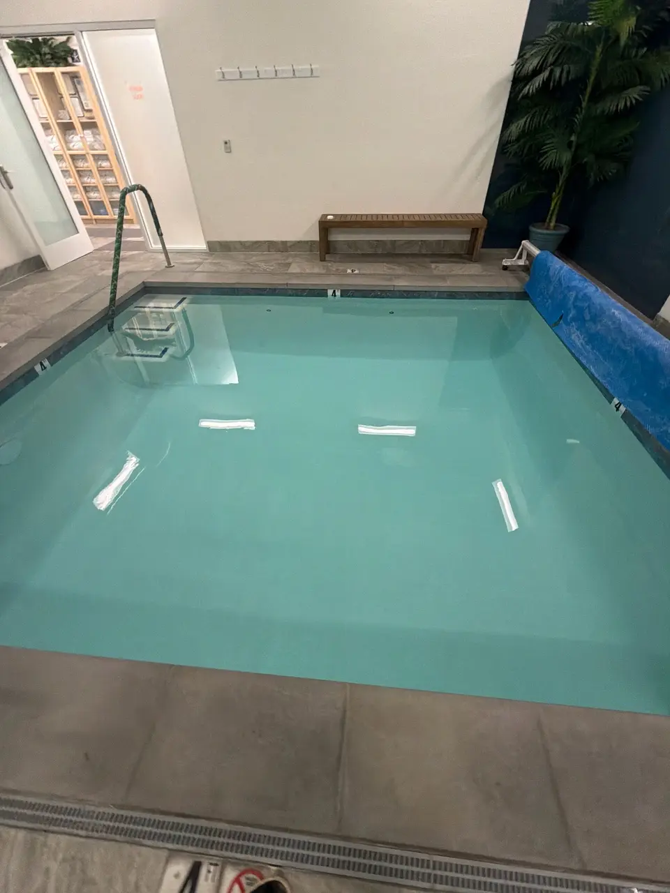 Clear turquoise water fills an indoor rectangular pool with an entry ladder, tiled deck, and poolside bench, after pool service in San Diego, CA